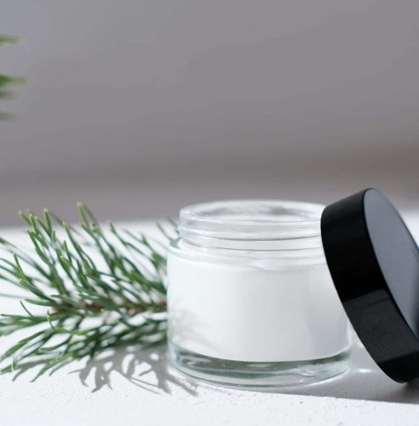 cold protection cream for dry skin. winter soothing and nourishing treatment for face care. white glass jar next to pine branches. Open cream container with cream inside